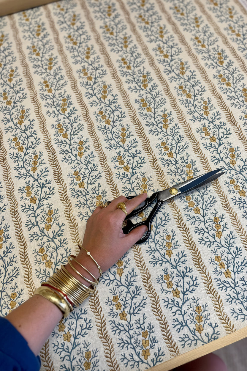 A hand wearing stacked gold bangles holds a pair of black-handled scissors over a cream-colored fabric with blue floral motifs and vertical tan stripes.