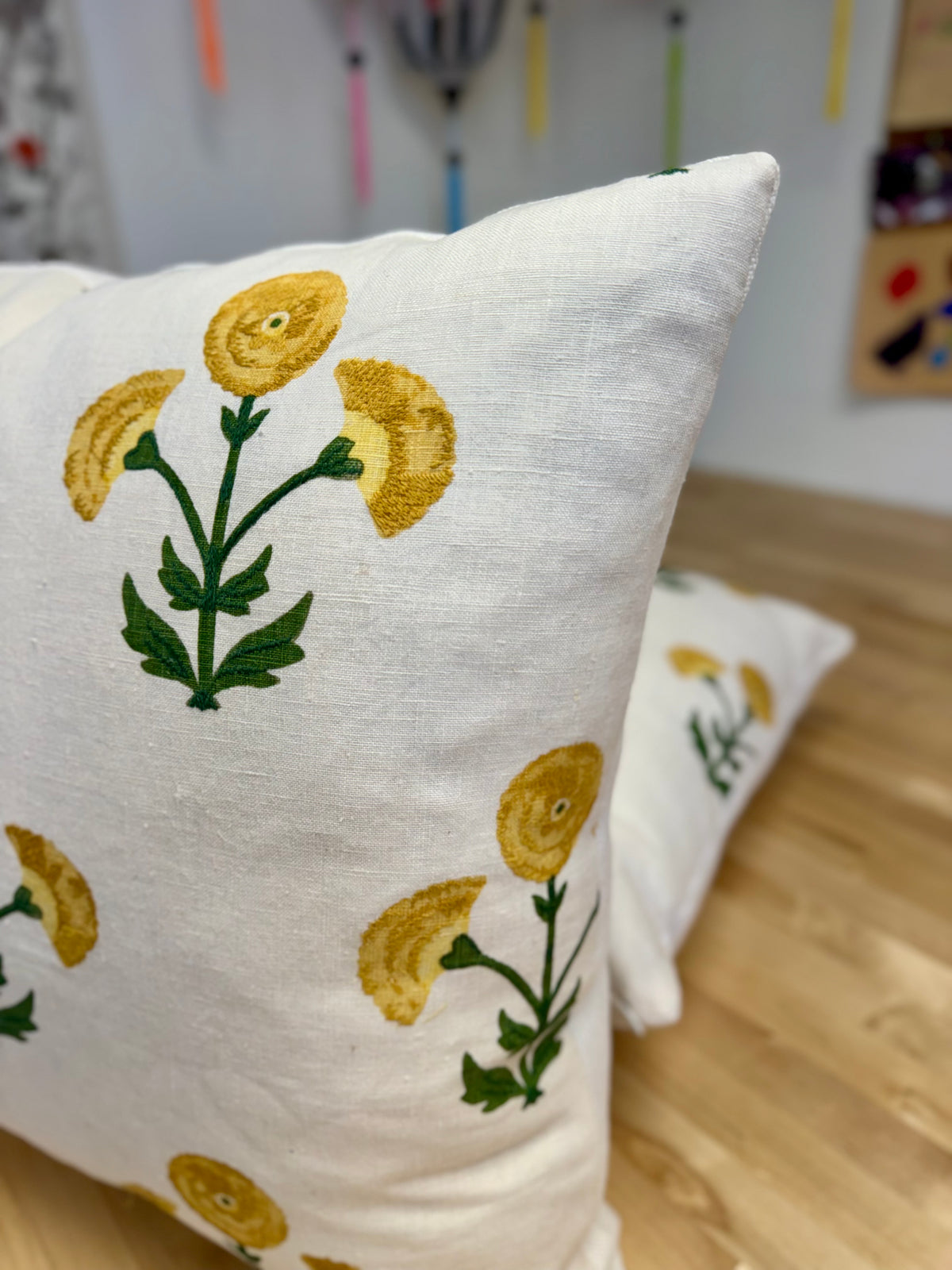 A close-up of a white throw pillow featuring a yellow and green floral pattern, with another matching pillow blurred in the background on a wooden floor.