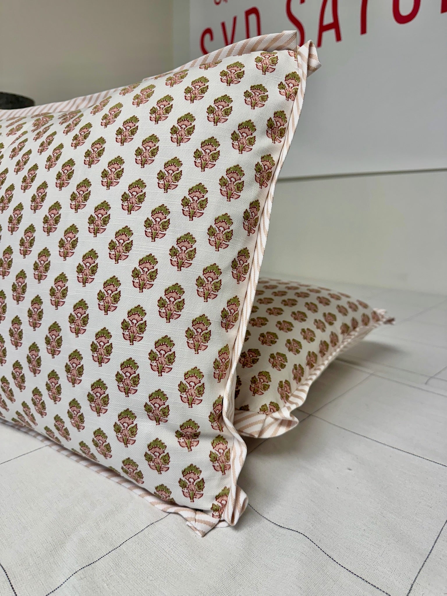 Two decorative throw pillows with a pink and green block-printed floral pattern on a cream-colored fabric, resting on a light-colored floor or bed.
