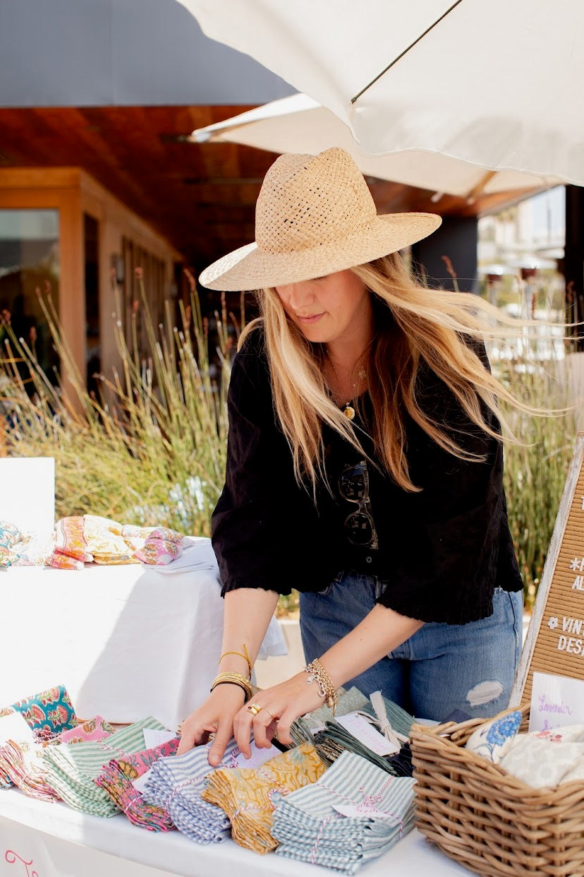 A woman wearing a straw hat and black top browses a table of colorful patterned cloth napkins at an outdoor market under a white umbrella.