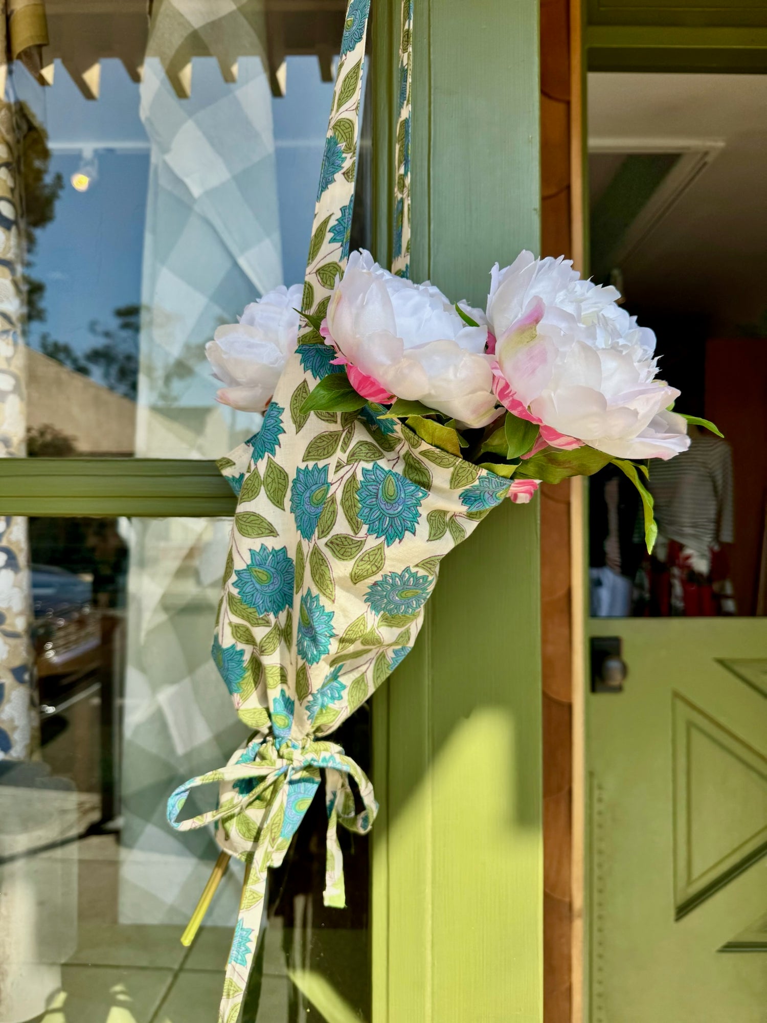 A fabric flower garland with white and pink peony-like flowers, tied with a matching floral ribbon, hanging on a green door frame in front of a window.