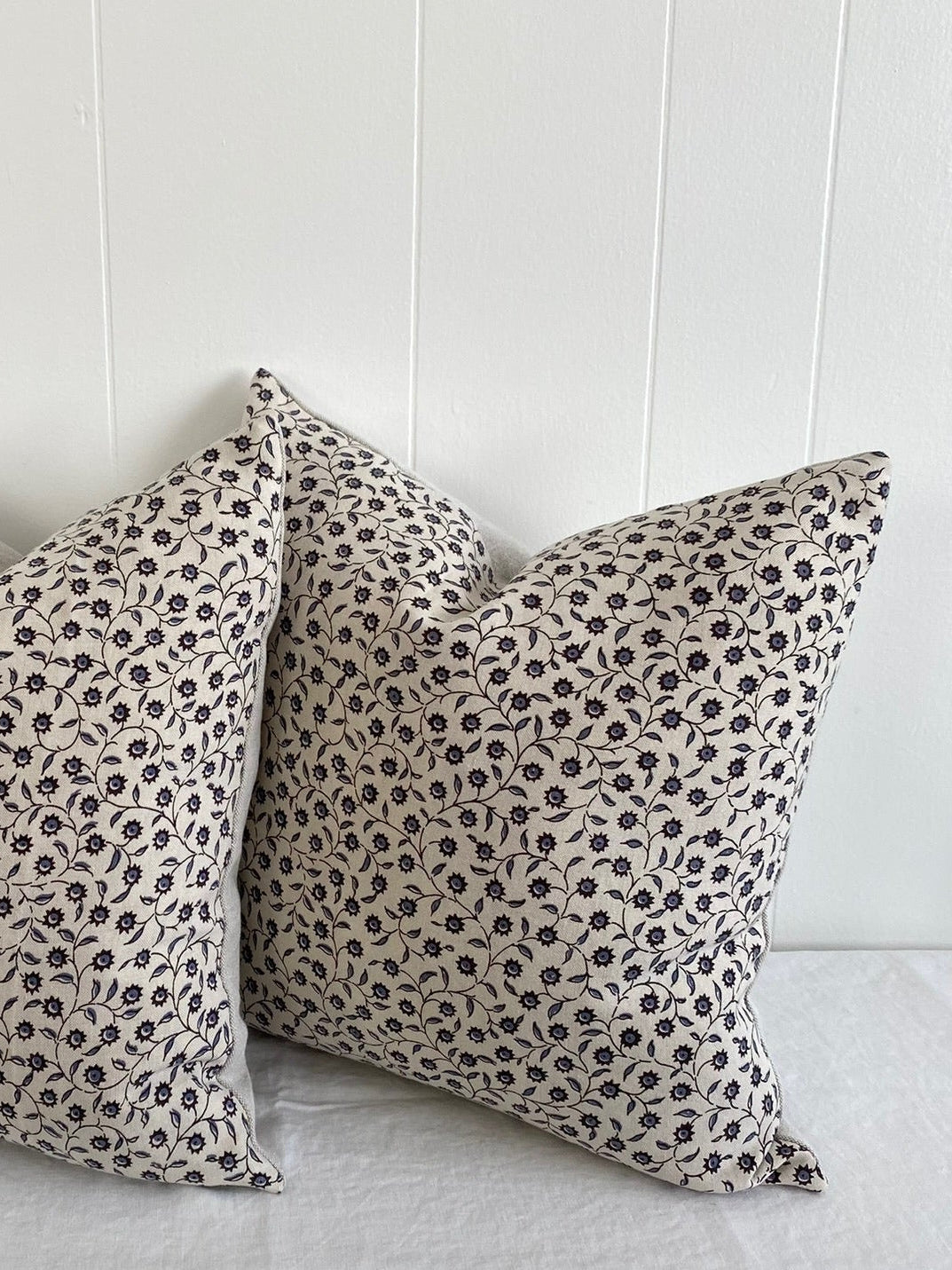 Two square throw pillows with a black leopard print on a warm beige background, resting on a white fabric-covered surface against a white paneled wall.