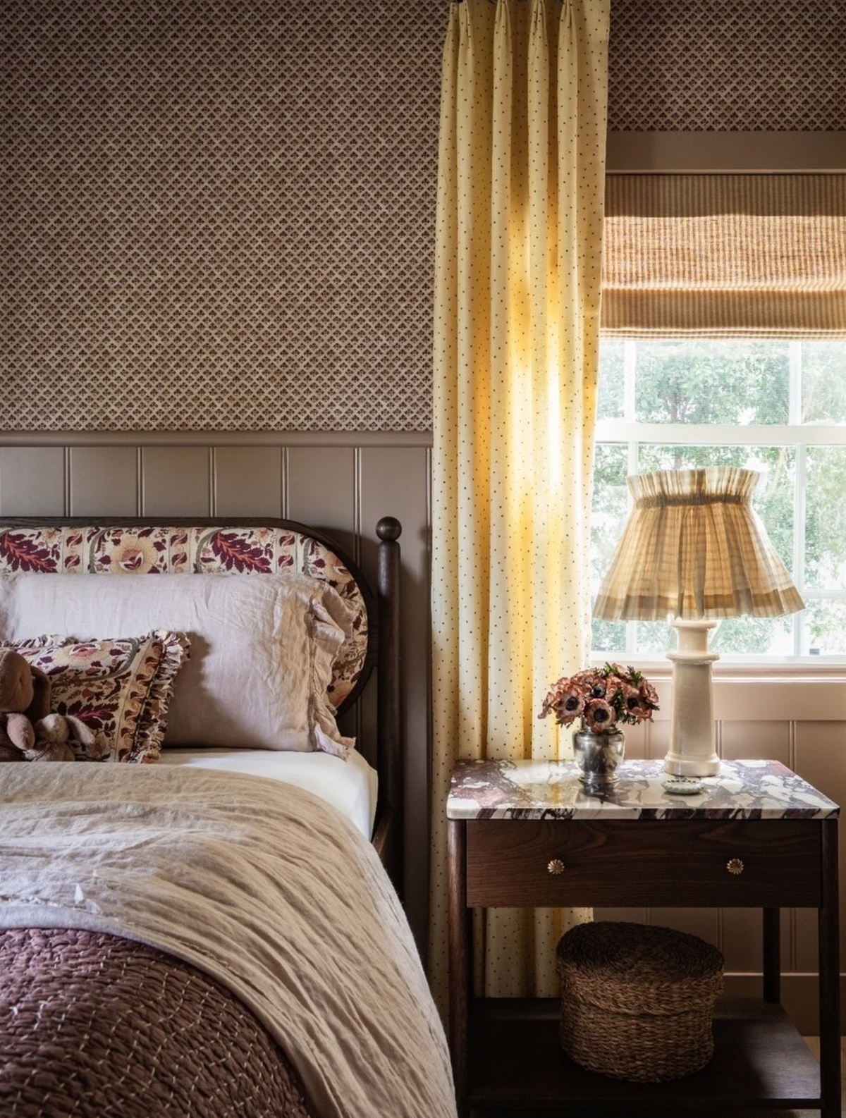 A cozy bedroom scene with a bed dressed in cream and brown textiles, patterned pillows, a dark wood headboard, a matching nightstand with a lamp and flowers, and yellow curtains framing a window.