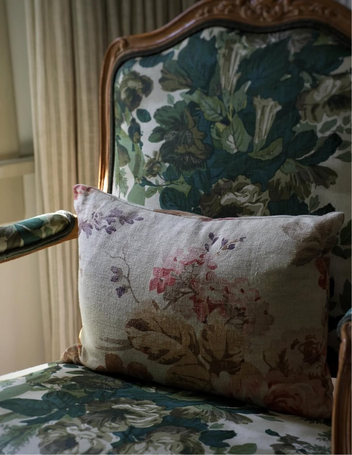 A square decorative throw pillow with a muted floral pattern in beige, pink, and brown, resting on an antique-style upholstered armchair with green and blue floral fabric.