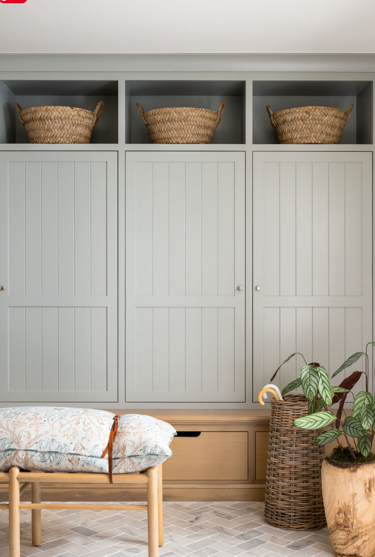 A styled interior scene showing a large built-in cabinet unit in a muted sage green color, with three open shelves at the top holding woven baskets, a bench with a patterned cushion in front, and potted plants on the floor.