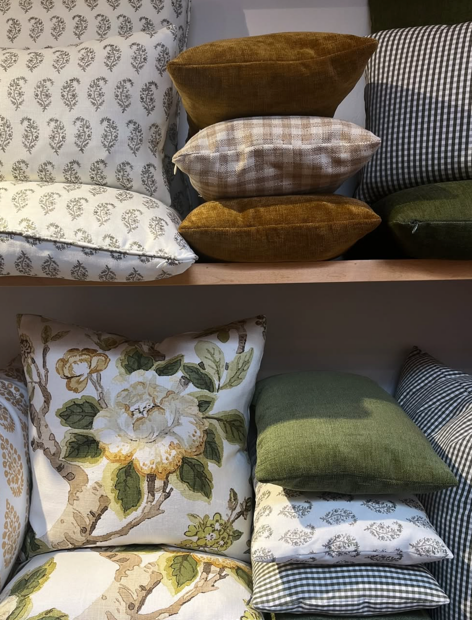 A collection of decorative throw pillows displayed on wooden shelves, featuring various patterns including floral, gingham, and plaid in muted earthy tones.