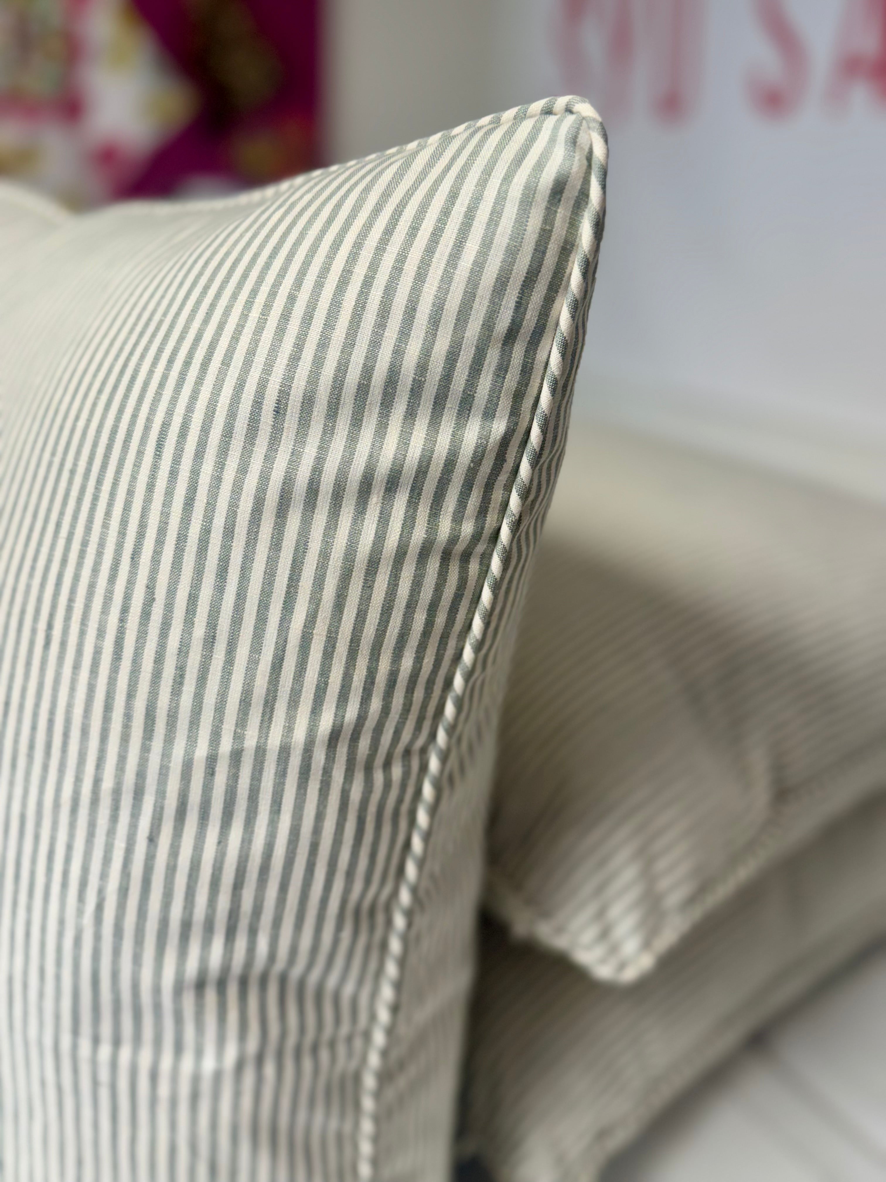 A close-up of a striped throw pillow in a light beige and gray fabric, showing its stitched edge and texture, with another matching pillow blurred in the background.
